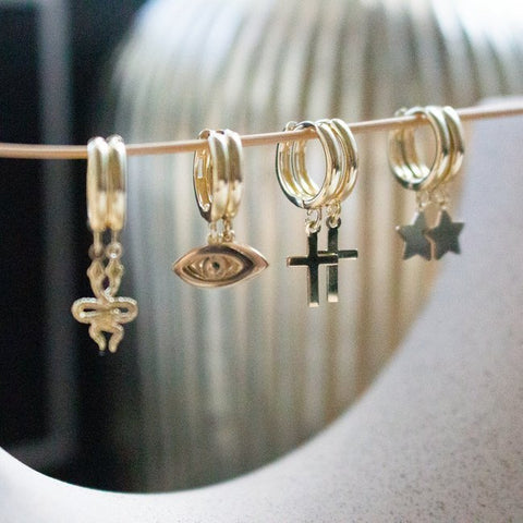 Set of gold hoop huggies earrings with various charms on a white surface - cross, evil eye, snake, star